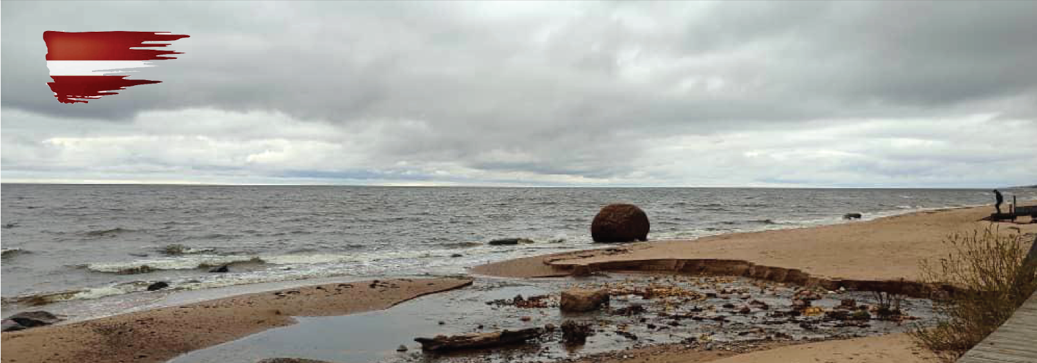 “Skats uz pelēku un mākoņainu jūras ainavu ar smilšainu krastu un lielu akmeni pie ūdens malas. Priekšplānā redzama neliela strauta ieplūde jūrā. Attēla augšējā kreisajā stūrī – sarkans un balts krāsu triepiens, kas atgādina Latvijas karogu.
